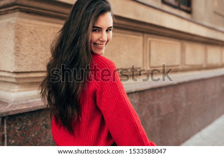 Outdoor rear view portrait of beautiful young brunette woman smiling, follow me on the city street. Pretty female model in trendy knitted red sweater walking in the town. Travel, people, lifestyle