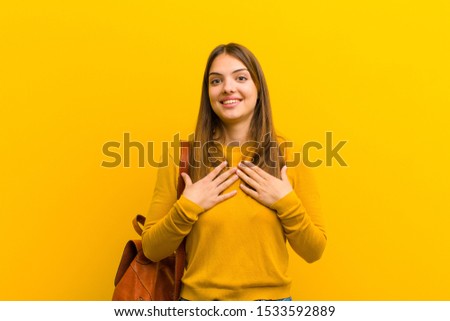 young pretty woman looking happy, surprised, proud and excited, pointing to self against orange background