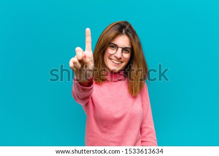 young pretty woman smiling proudly and confidently making number one pose triumphantly, feeling like a leader against blue background