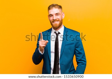 young red head businessman smiling and looking friendly, showing number one or first with hand forward, counting down against orange background
