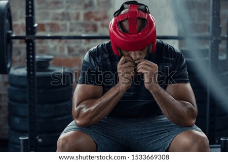 African american man in red boxing head gear putting gumshield. Determined boxer in sportswear sitting near boxing ring. Young man preparing for boxe practice with protective helmet. Royalty-Free Stock Photo #1533669308