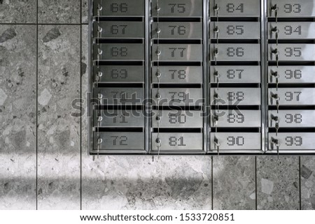 Lot of numbered metallic postboxes with inserted keys are hanging on the textured gray wall indoors. Sunlight falls down on them. Closeup horizontal photo.