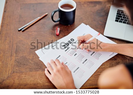 Stock photo of female hands holding pen and making notes on calendar page for year 2020. Over shoulder view businesswoman doing time planning for the next year. Concept of scheduling