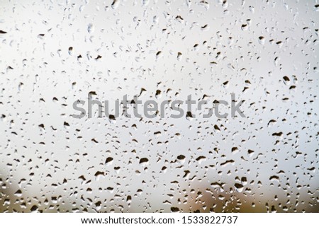 Pattern of dripping raindrops on a window or any other transparent surface on a cloudy autumn day, the texture of water droplets close-up on the glass with a blurred cityscape on the background.