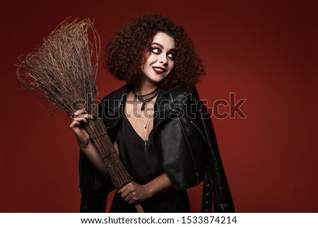 Image of beautiful witch girl in black dress with halloween makeup holding broom isolated over red wall