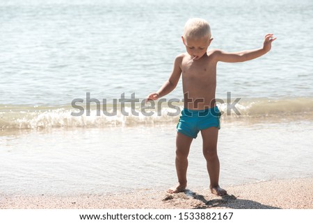 little blond boy in blue shorts with a lollipop in his hands smiles and shows various facial expressions by the sea
