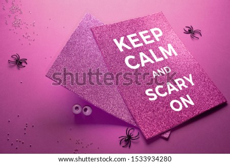 Creative purple Halloween background with spiders, chocolate eyes and two glittering cards on fuchsia colored paper. Text "Keep calm and scary on" on the top card.