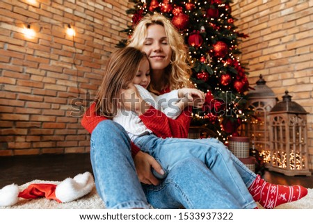 Christmas. Childhood. Home. Little girl and her mom are hugging and smiling while sitting near the Xmas tree