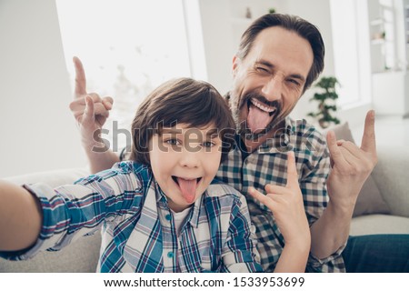 Hey there followers! Close up photo of positive cheerful handsome guy dad making hard punk symbol and his little son taking selfie on smartphone showing tongue in room indoors Royalty-Free Stock Photo #1533953699