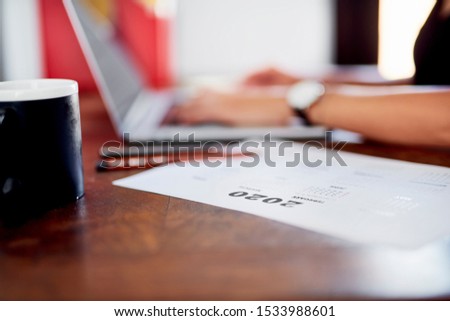 Stock photo of calendar for year 2020 lying on table with blurred office worker typing on laptop on background, focus on numbers. Crop girl using gadget at workplace. Concept of planning