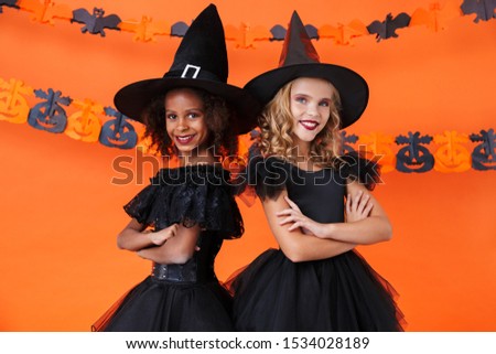 Image of nice witch girls in black halloween costumes smiling and standing together isolated over orange pumpkin wall