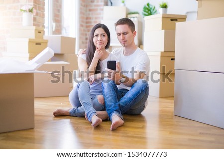 Beautiful couple sitting on the floor using smartphone at new home around cardboard boxes serious face thinking about question, very confused idea