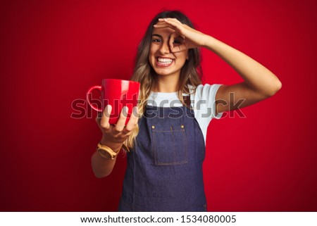 Young beautiful barista woman wearing apron over red isolated background with happy face smiling doing ok sign with hand on eye looking through fingers