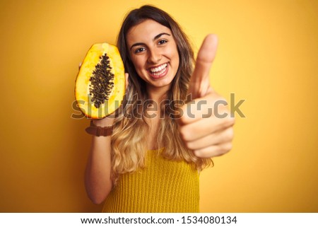 Young beautiful woman holding papaya over yellow isolated background happy with big smile doing ok sign, thumb up with fingers, excellent sign