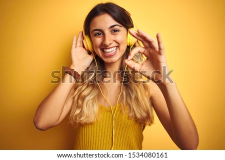Young beautiful woman wearing headphones over yellow isolated background doing ok sign with fingers, excellent symbol