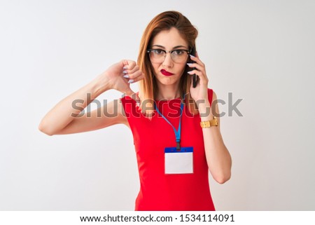 Redhead businesswoman wearing id card talking on smartphone over isolated white background with angry face, negative sign showing dislike with thumbs down, rejection concept