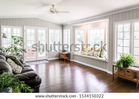 Large and open living room den sun room with windows on two sides and lots of natural light flowing in. There is a window seat on one side and a leather couch and plant on the other. Royalty-Free Stock Photo #1534150322