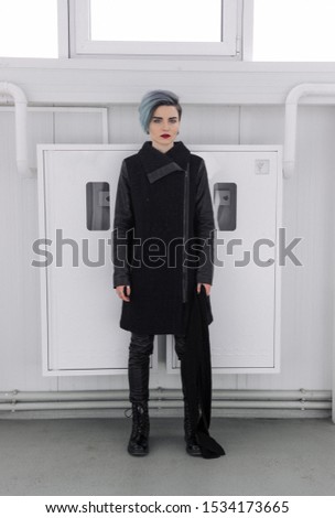 Beautiful girl with short blue fashionable hairstyle in black coat posing on the light background. Head turning right, hands along the body. Full-length photo.