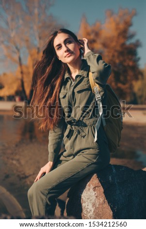 Tourist woman with backpack sitting on stone near river in autumn day