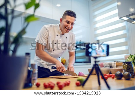 Smiling caucasian chef in uniform standing in kitchen and cutting onion while filming himself for blog. On kitchen counter are vegetables and spices. Royalty-Free Stock Photo #1534215023