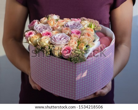 flowers and sweets in a heart-shaped box in the hands of a girl in the sun as a gift. purple one-headed rose, rose Bush rose, beige Bush rose, Sedum, pink macaroni, close-up with blurred background