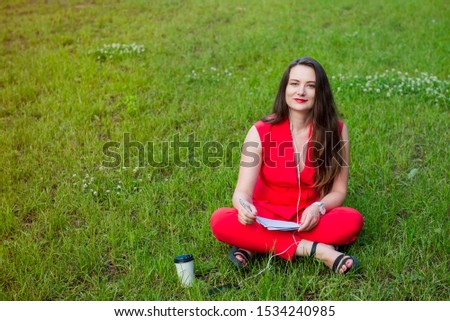 Portrait of young smiling business woman in red suit in earphones listening to music, doing online learning cources and writing in notebook while sitting on the green grass lawn in park. Copy space
