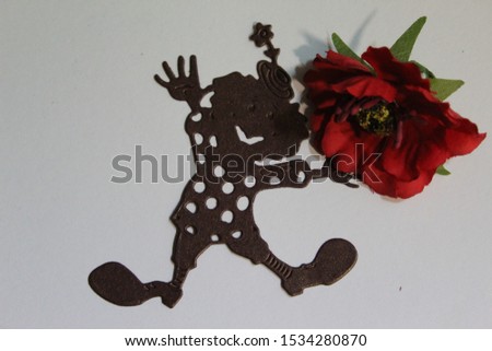 Photo of a brown, paper clown, with a red poppy flower, on a white background.

