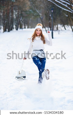 Beautiful young woman weared in white sweater and hat with ice skates on the hands having fun in winter snowy park.