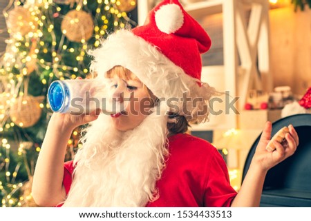 Greeting card background. Cheerful little Santa Claus holding glass with milk and cookie with fireplace and Christmas Tree in the background