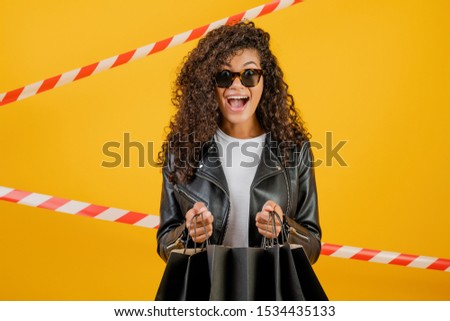 trendy young black woman wearing jacket with paper shopping bags isolated over yellow with signal tape