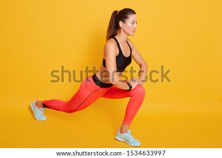 Indoor shot of attractive brunette young slim woman wearing stylish sports clothing stretching in gym, young female doing warmup stretching workout, isolatedover yellow background. Fitness concept.