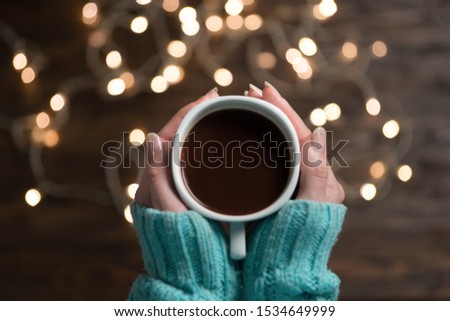 Night composition with woman hands holding cup of coffee. Warm garland. Autumn home comfort.