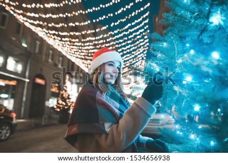 Christmas night portrait of a girl near a Christmas tree on the background of a street decorated with a garland, looks at the lights and touches the branches of the Christmas tree.new Year
