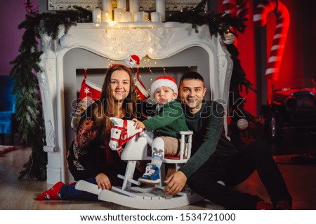 The family sits under a large white fireplace decorated
red socks and Christmas lights. Mom and dad rock baby on wooden horse against fireplace background. The concept of Christmas, New Year, gifts.