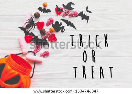 Trick or treat text, happy Halloween sign. Halloween candy spilled from jack o lantern bucket with skulls, black bats, ghost, spider on white wooden background, flat lay. 