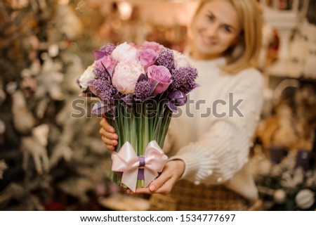 Smiling young woman holding a bouquet of tender pink and purple color flowers with green stalks in the blurred background of flower shop