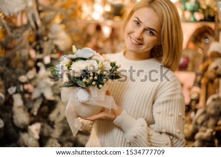 Woman holding a flower pot with white flowers decorated wuth green leafs and fir-tree branches in the blurred background of flower shop