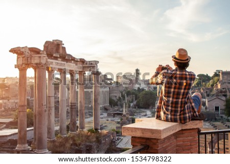 Young man tourist sitting at Roman Forum at sunrise taking picture with vintage camera. Historical imperial Foro Romano from panoramic point of view, Italy