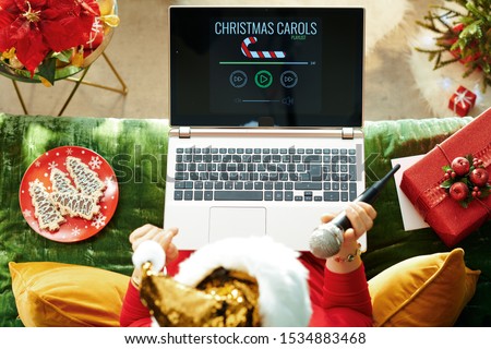 Holiday season. Upper view of stylish woman in sparkle gold sequin santa hat sitting on divan in the modern living room at Christmas and signing while listening Christmas songs on a laptop.