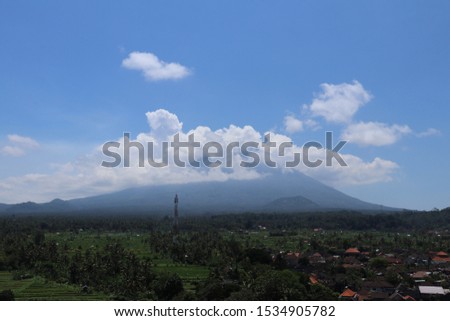 Aerial view of majestic Gunung Agung volcano on tropical Bali island in Indonesia. Rice fields and palm trees in valley below mountain. The top of the volcano partially shrouded in thick white clouds.