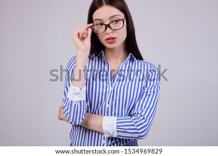 Cute business girl with brown hair, full pink lips posing on the background. She is wearing a white and blue striped shirt and black computer glasses, which she holds with her right hand. Looks side.