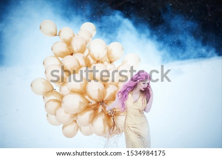 Mysterious art portrait of beautiful woman with pink hair in golden fashionable long dress with hairstyle and make up against the background of snowy winter park. Girl with bunch of golden balloons 