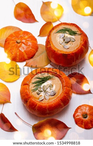 Fresh creamy orange pumpkin soup in baked pumpkins with green dill and pumpkin seeds on white wooden background with yellow and red autumn leaves and bokeh lights. Top view