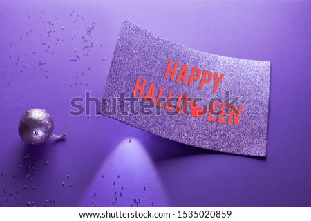 Creative purple Halloween background with glittering pumpkin painted metallic on neon paper. Spotlight shining from underneath glittering paper card with text "Happy Halloween".