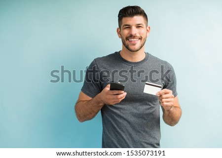 Happy smart young male millennial holding credit card and mobile phone against turquoise background