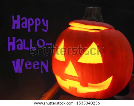 Happy Halloween, purple violet inscription on a black background, near the orange, carved pumpkin, with shining eyes. Jack o lantern or smiling Halloween Jack. Greeting card. Specially blurred image.