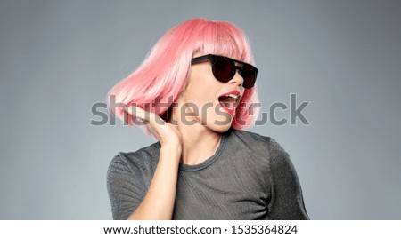 style, fashion and people concept - happy laughing young woman in pink wig and black sunglasses dancing over grey background