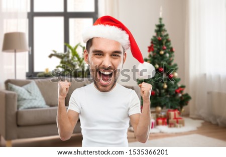 emotion, expression and winter holiday concept - young man in santa hat celebrating victory and screaming over christmas tree on home background (funny cartoon style character with big head)