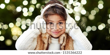 christmas, season and holiday concept - happy little girl wearing earmuffs over festive lights on dark green background