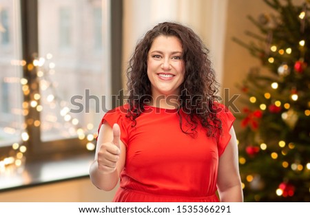 winter holidays, people and gesture concept - happy woman in red dress showing thumbs up over christmas tree lights on home background